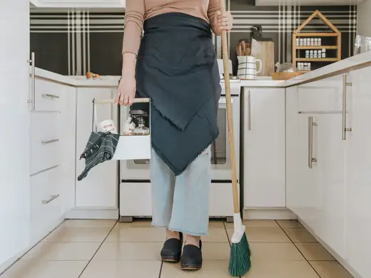 Person in an apron holding a broom and cleaning caddy in a white kitchen