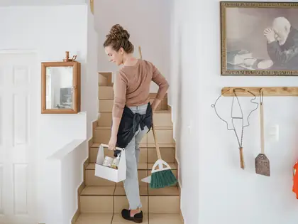 Woman carrying a cleaning caddy and broom up tiled stairs